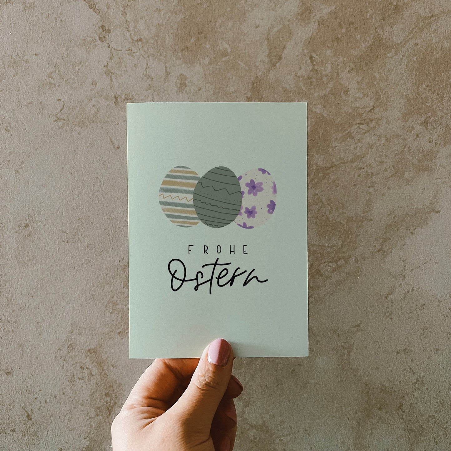 Postkarte im Hochformat, Abgebildet sind drei bunte Eier in Pastellfarben. Der Hintergrund ist Pastellgrün. Auf der Karte steht ein Lettern mit dem Text Frohe Ostern
