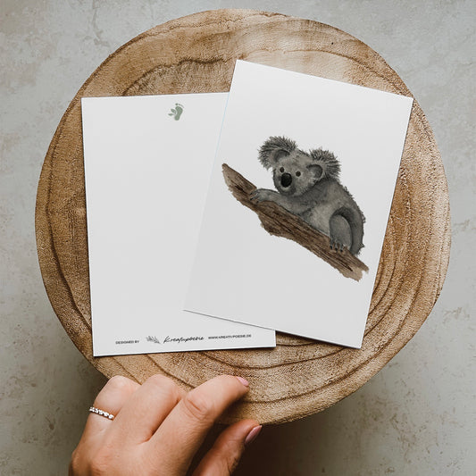 Postkarte DIN A6, Hochformat, gedruckt in Deutschland. Auf dieser Karte befindet sich ein junger Koala, welcher sich gemütlich einen Baumstamm klammert.
