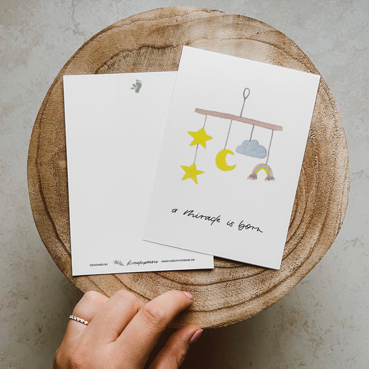 Postkarte im Hochformat. Anlass: Geburt. In der Mitte ist ein aquarelliertes Mobile abgebildet. Unterhalb dessen befindet sich der von Hand geschriebene Schriftzug ,,a miracle is born''. Die Karte ist in den Tönen gelb, blau und braun geahalten.