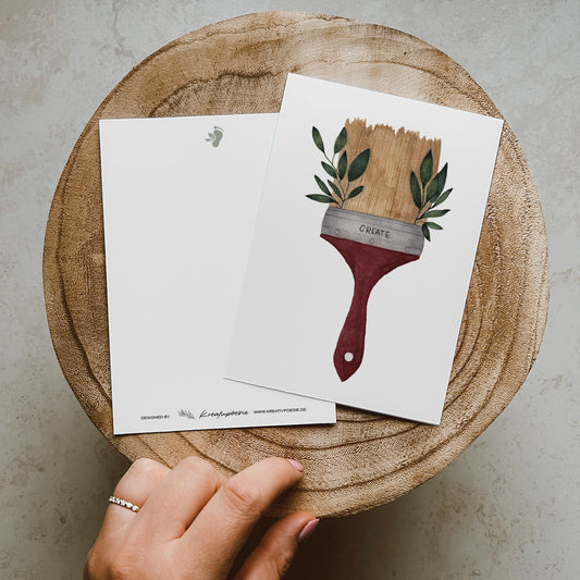Postkarte im Hochformat. Malerpinsel mit grünen Blättern an der Seite und Lettering ,,create''. Der Pinsel ist beige und der Stiel rot.
