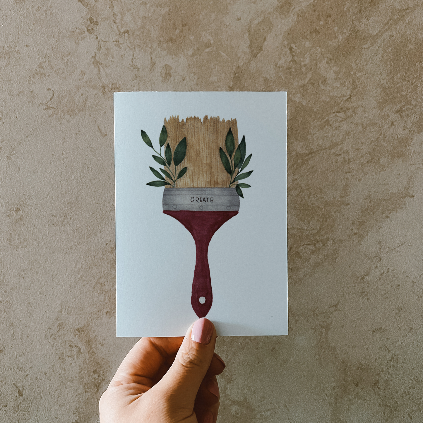 Postkarte im Hochformat. Malerpinsel mit grünen Blättern an der Seite und Lettering ,,create''. Der Pinsel ist beige und der Stiel rot.