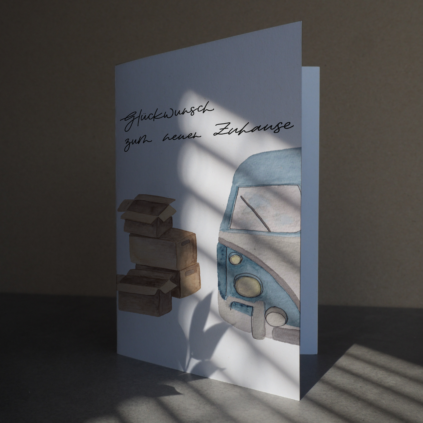 Klappkarte mit blauem Bulli, neben welchem vier gestapelte Umzugskartons stehen. Lettering: ,,Glückwunsch zum neuen Zuhause''