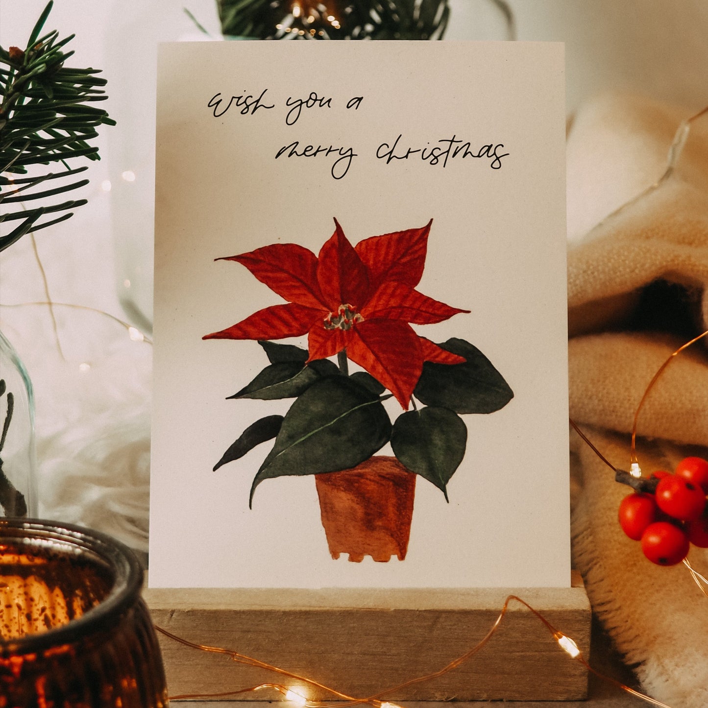 Postkarte im Hochformat. Auf ihr befindet sich ein roter Weihnachtsstern, der in einem braunen Tontopf steht. Oberhalb dessen steht mein ahandgeschriebener Schriftzug »Wish you a Merry Christmas«.