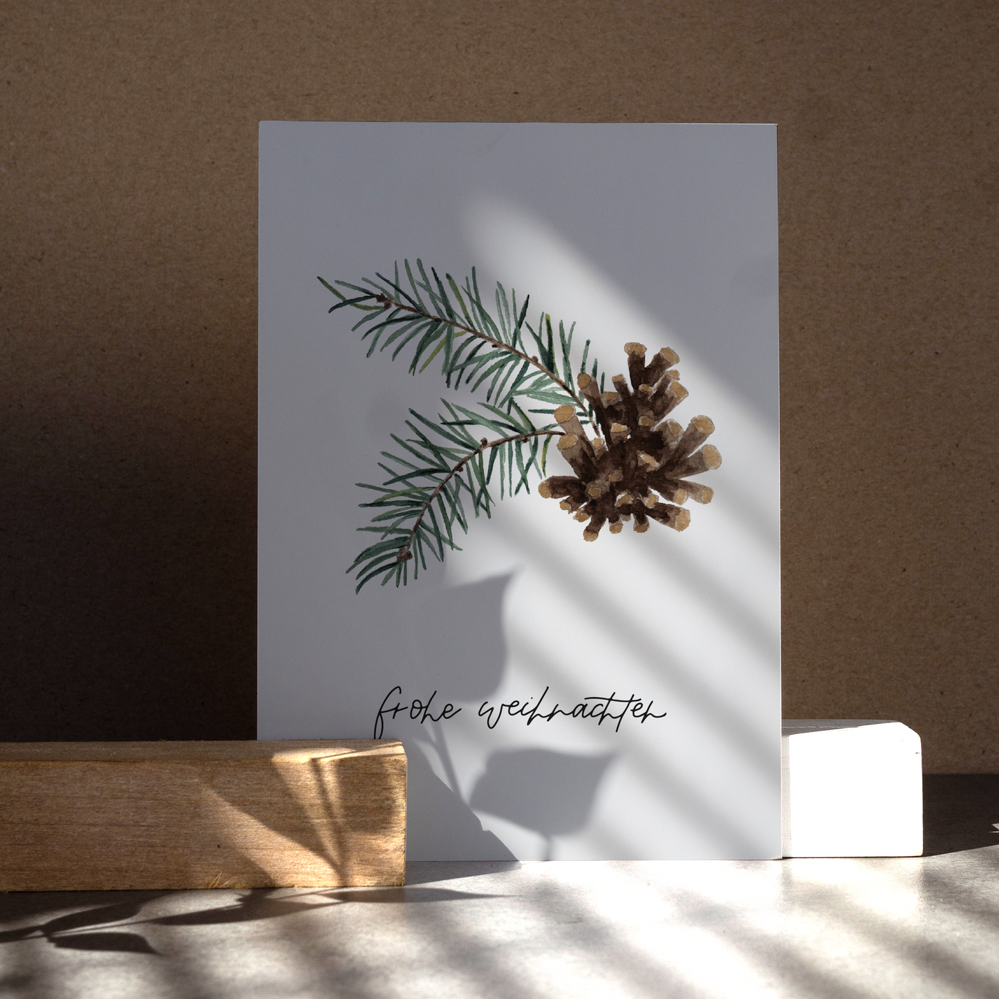 Postkarte im Hochformat. Auf ihr befindet sich ein Tannenzapfen, welcher umgeben ist von zwei Tannenzweigen. Unter ihm befindet sich mein handgeschriebener Schriftzug »frohe Weihnachten«.