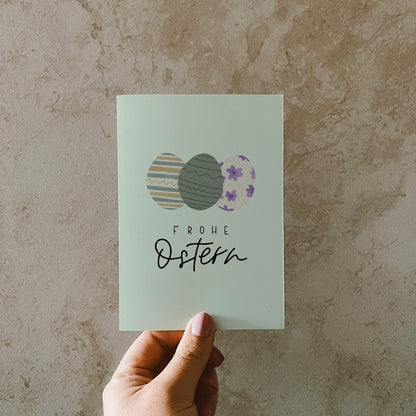 Postkarte im Hochformat, Abgebildet sind drei bunte Eier in Pastellfarben. Der Hintergrund ist Pastellgrün. Auf der Karte steht ein Lettern mit dem Text Frohe Ostern