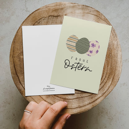Postkarte im Hochformat, Abgebildet sind drei bunte Eier in Pastellfarben. Der Hintergrund ist Pastellgrün. Auf der Karte steht ein Lettern mit dem Text Frohe Ostern