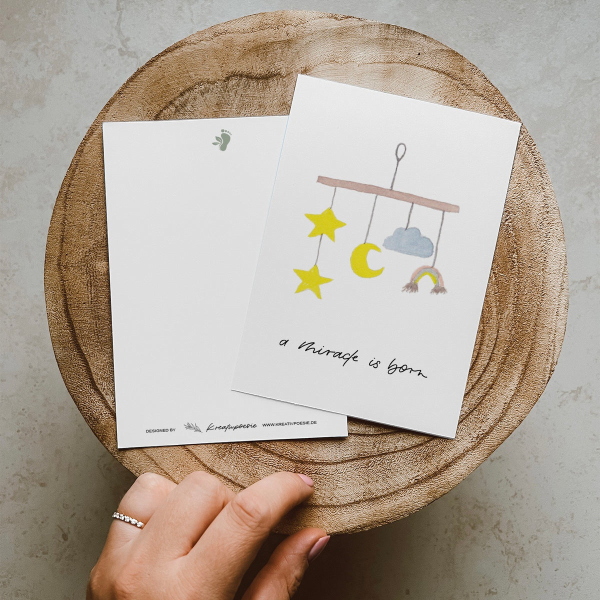 Postkarte im Hochformat. Anlass: Geburt. In der Mitte ist ein aquarelliertes Mobile abgebildet. Unterhalb dessen befindet sich der von Hand geschriebene Schriftzug ,,a miracle is born''. Die Karte ist in den Tönen gelb, blau und braun geahalten.