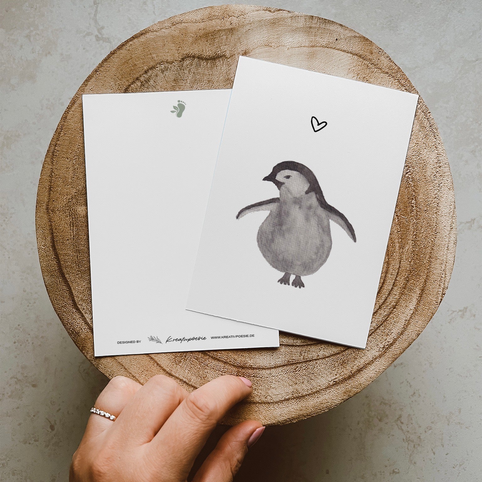 Grau schwarzer Pinguin mit abstehenden Flügeln, nach links schauend mit einem Herz über ihm. Gedruckt auf Recyclingpapier in Deutschland.