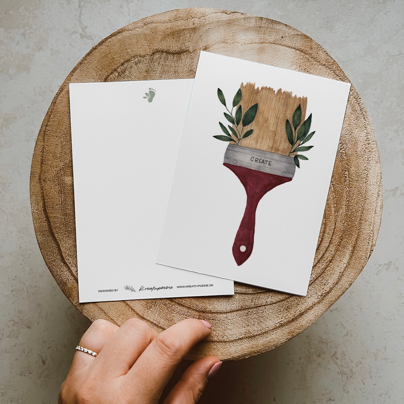 Postkarte im Hochformat. Malerpinsel mit grünen Blättern an der Seite und Lettering ,,create''. Der Pinsel ist beige und der Stiel rot.
