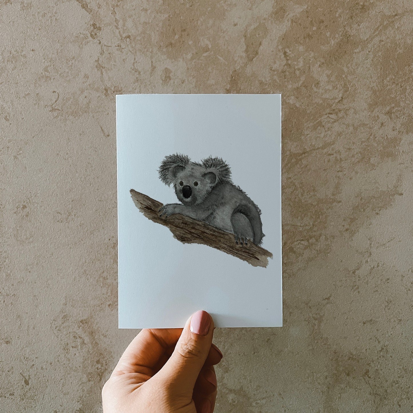 Postkarte DIN A6, Hochformat, gedruckt in Deutschland. Auf dieser Karte befindet sich ein junger Koala, welcher sich gemütlich einen Baumstamm klammert.