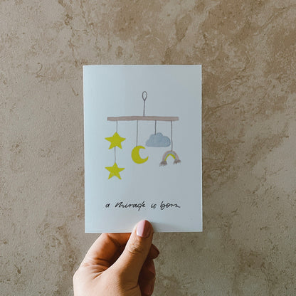 Postkarte im Hochformat. Anlass: Geburt. In der Mitte ist ein aquarelliertes Mobile abgebildet. Unterhalb dessen befindet sich der von Hand geschriebene Schriftzug ,,a miracle is born''. Die Karte ist in den Tönen gelb, blau und braun geahalten.
