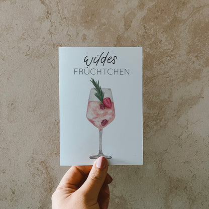 Postkarte DIN A6, Hochformat, gedruckt in Deutschland. Auf dieser Karte befindet sich ein Weinglas mit Früchten und ein rosa gefärbtes Getränk, welches an Lille Wild Berry erinnern soll. Über ihm befindet sich ein Lettering ,,wildes Früchtchen''.