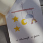 Postkarte im Hochformat. Nahansicht. Geburtskarte, in der Mitte ist ein aquarelliertes Mobile abgebildet. Unterhalb dessen befindet sich der von Hand geschriebene Schriftzug ,,a miracle is born''. Die Karte ist in den Tönen gelb, blau und braun geahalten.