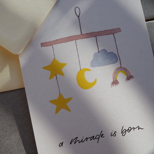 Postkarte im Hochformat. Nahansicht. Geburtskarte, in der Mitte ist ein aquarelliertes Mobile abgebildet. Unterhalb dessen befindet sich der von Hand geschriebene Schriftzug ,,a miracle is born''. Die Karte ist in den Tönen gelb, blau und braun geahalten.