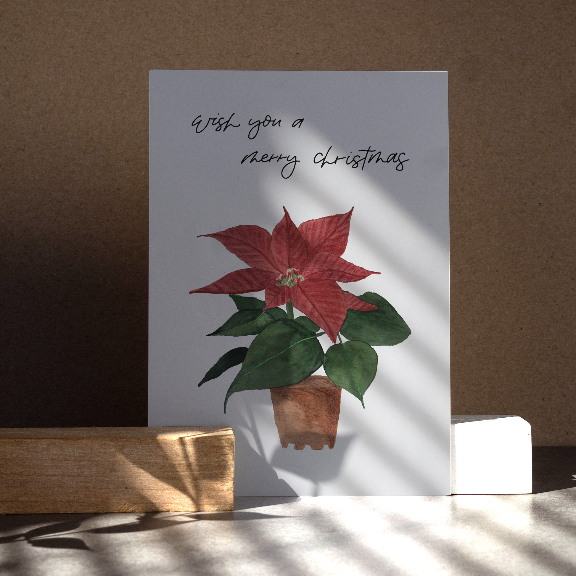 Postkarte im Hochformat. Auf ihr befindet sich ein roter Weihnachtsstern, der in einem braunen Tontopf steht. Oberhalb dessen steht mein ahandgeschriebener Schriftzug »Wish you a Merry Christmas«.