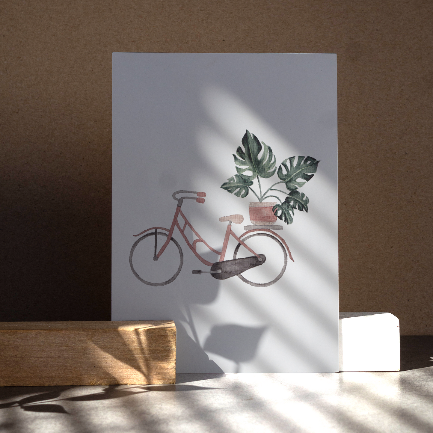 Postkarte DIN A6, Hochformat, gedruckt in Deutschland. Eine Monstera im rosa-beigen Blumentopf, welcher sich auf einem Gepäckträger eines rosa Fahrrads befindet.